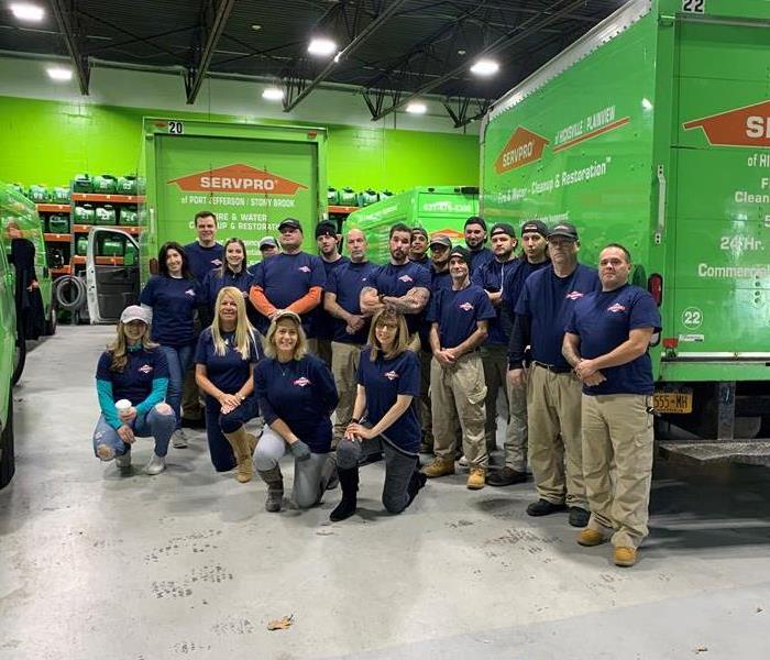 A large group of SERVPRO employees standing in a green garage with equipment and SERVPRO vehicles.  